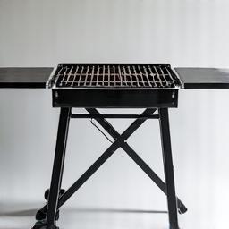 A portable, foldable grill stand with a built-in side table, showcasing its compact storage and robust setup.